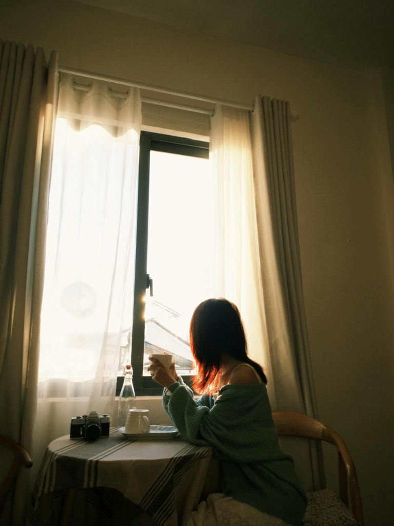 A woman sitting by a window with a cup of tea, reflecting quietly in soft morning light.